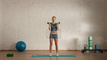 Photo for Personal sport classes at home online. Blonde female in sportswear doing exercises. Healthcare creative advertisement concept. Woman fitness coach in the room doing biceps with dumbbells. - Royalty Free Image