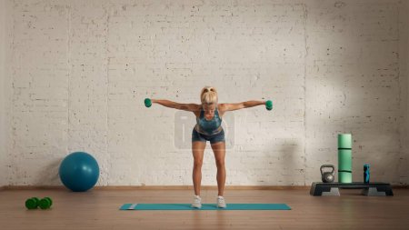 Photo for Personal sport classes at home online. Blonde female in sportswear doing exercises. Healthcare creative advertisement concept. Woman fitness coach in the room doing dumbbells half bend swing. - Royalty Free Image