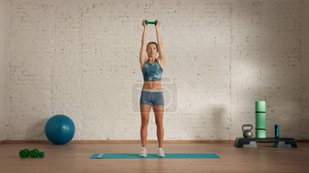 Photo for Personal sport classes at home online. Blonde female in sportswear doing exercises. Healthcare creative advertisement concept. Woman fitness coach in the room holding dumbbell over head. up. - Royalty Free Image