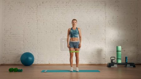 Photo for Personal sport classes at home online. Blonde female in sportswear doing exercises. Healthcare creative advertisement concept. Woman fitness coach in the room standing with rubber band over thighs. - Royalty Free Image