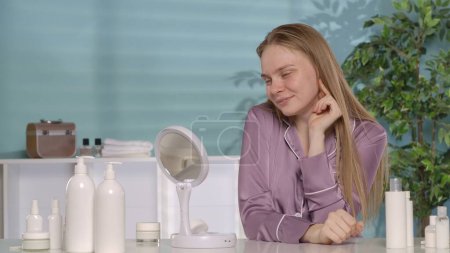 Photo for Portrait of a woman sitting at a dressing table in front of a mirror. Woman without makeup and in pajamas is satisfied with the cleanliness and smoothness of the skin of the face - Royalty Free Image