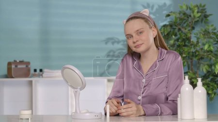 Photo for Portrait of a joyful woman sitting at a dressing table in front of a mirror. A woman without makeup, wearing a headband and pajamas is satisfied with the cleanliness and smoothness of her facial skin - Royalty Free Image