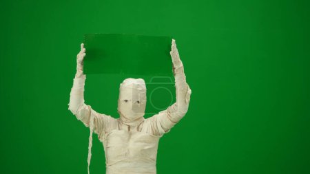 Pantalla verde aislada croma clave foto capturar una momia espeluznante sosteniendo un cartel de pantalla verde. Prepárate para tu clip de promoción o anuncio. Tamaño medio.