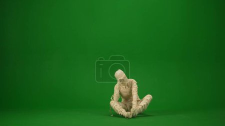 Foto de Una momia cansada o frustrada se sienta en el suelo y sostiene su cabeza. Tecla de croma aislada de pantalla verde. Mock up, espacio de trabajo, publicidad. Longitud total. Halloween vacaciones. - Imagen libre de derechos
