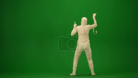 Photo for A mummy wrapped in bandages poses or dances with his arms raised. Green screen isolated chroma key. Mock up, workspace, advertisement. Full length. Back view. Halloween holidays. - Royalty Free Image