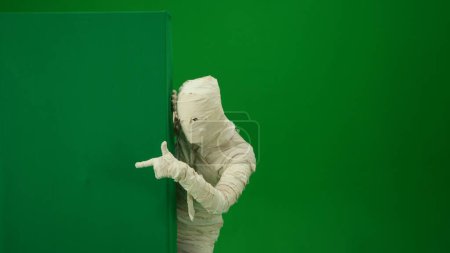 Photo for Green screen isolated chroma key photo capturing a mummy peeking from behind a green screen banner box and pointing at it. Medium size. Mock up, workspace for your promotion clip or advertisement. - Royalty Free Image