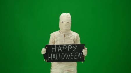 Photo for Green screen isolated chroma key photo capturing a mummy holding a chalkboard with happy halloween on it. Mock up for your promotion clip or advertisement. Medium size. - Royalty Free Image