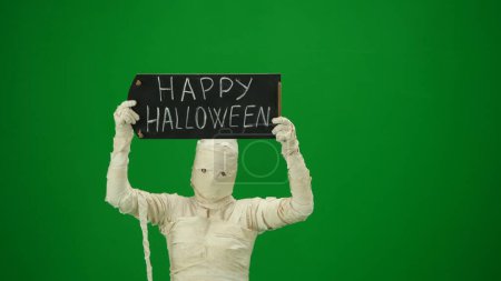 Téléchargez les photos : Écran vert isolé chroma photo clé capturant une momie tenant un tableau avec halloween heureux sur elle. Maquette pour votre clip de promotion ou publicité. Taille moyenne. - en image libre de droit