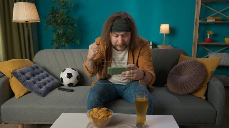 Photo for In the picture, a middle aged man is sitting on a sofa in the living room. He is holding a phone and looking gesticulating on it something. Next to him lies a ball and beer with chips. He is attentive - Royalty Free Image
