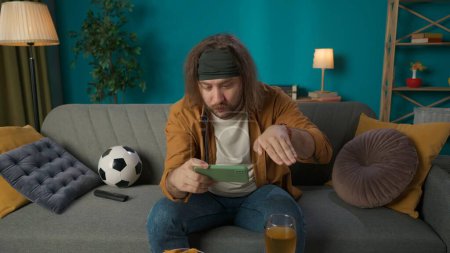 Photo for In the picture, a middle aged man is sitting on a sofa in the living room. He is holding a phone and looking gesticulating on it something. Next to him lies a ball and beer with chips. He is attentive - Royalty Free Image