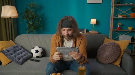 Photo for In the shot, a middle aged man is sitting on a sofa in a room. He is holding a tablet and is watching something in it. Next to the ball there is a beer and chips on the table. It is concentrated. - Royalty Free Image