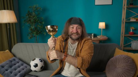 Photo for A football fan a middle aged man is sitting on a couch. Next to him is a ball. In his hands he holds a gold cup, joyfully clutching it. Showing the excitement of the football team winning. Medium shot - Royalty Free Image