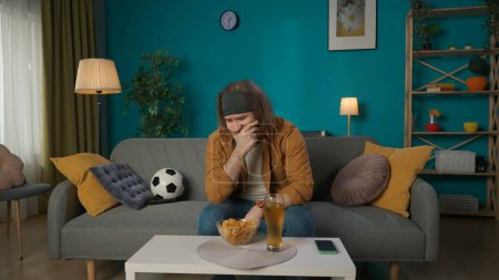 Photo for In the picture, a middle aged man is sitting on a sofa in a room. Next to the soccer ball is a beer and chips. He looks away with his hand over his face and plays a football fan, looks upset, sad - Royalty Free Image
