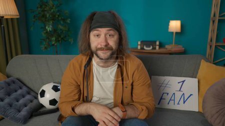 Photo for In a close up shot, a middle aged man sits on a couch in a room and looks at the camera. A soccer ball lies nearby. He portrays a soccer fan, showing excitement, concentrated, in anticipation. - Royalty Free Image