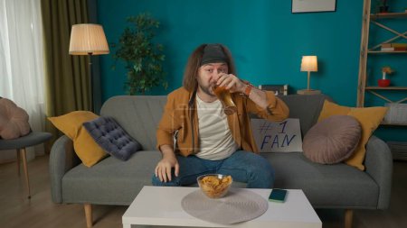 Photo for A middle aged man is sitting on a sofa. Next to him is a fan tablet, on the table there are beer, chips and a phone. He looks at the camera and drinks beer. Portrays a football fan watching a match. - Royalty Free Image