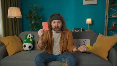 Photo for Middle aged man sitting on a couch next to a ball and a fans hat. He looks into the camera and holds out a red card and holds up a yellow card. Imitates a fan who is refereeing a soccer match. - Royalty Free Image