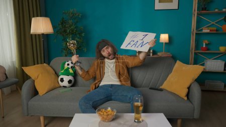 Photo for A middle aged man sits on a couch in a room. He is looking at the camera, yelling and holding a sign and a trophy. Imitates a soccer fan who supports the soccer team. Hes excited, waiting. Medium shot - Royalty Free Image