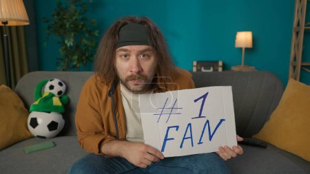 Photo for In the shot, a middle aged man is sitting on a couch. Hes holding a fan sign. He s looking at the camera to watch a football game. He is a fan and supports the team. - Royalty Free Image