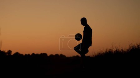 Téléchargez les photos : Photo pleine longueur capturant une silhouette d'un adolescent, jeune homme jouant avec une balle sur un terrain au coucher du soleil. Il donne un coup de pied dans les airs. Exercice, football et autres sports, jeunesse. - en image libre de droit