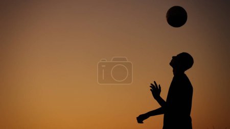 Téléchargez les photos : Photo de taille moyenne capturant une silhouette d'un adolescent, jeune homme jouant avec une balle à l'extérieur au coucher du soleil. Il lance la balle en l'air. Exercice, football et autres sports, jeunesse. - en image libre de droit