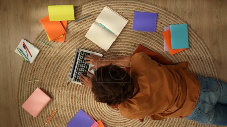 Photo for Top view capturing a young man laying on the floor and studying, working on a laptop and taking notes, surrounded with colorful books. College and university, homeschooling, online, education. - Royalty Free Image