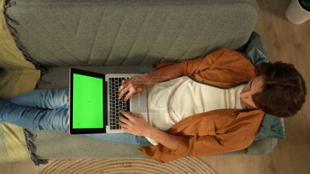 Téléchargez les photos : Vue de dessus capturant un jeune homme allongé sur le canapé, regardant l'écran de l'ordinateur portable avec une zone publicitaire, un espace de travail maquillé dessus. Loisirs, achats en ligne, réseaux sociaux et communication. - en image libre de droit