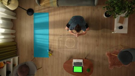 Photo for Top view capturing a young man doing squats in front of the laptop with a workspace mock up on it, as if he is watching a video. . Indoor activities. Healthy and active lifestyle, morning routine. - Royalty Free Image