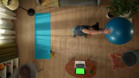 Photo for Top view capturing a young man doing exercises using an exercise ball in front of the laptop with a workspace mock up on it. Indoor activities. Healthy and active lifestyle, morning routine. - Royalty Free Image