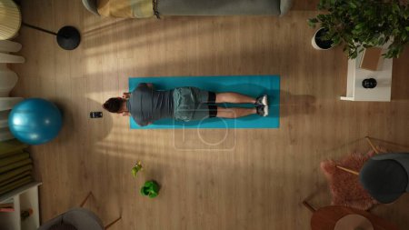 Photo for Top view capturing a young man doing his morning exercise, standing in a plank with a timer on his smartphone. Indoor activities. Healthy and active lifestyle, morning routine, achieving the goals. - Royalty Free Image