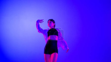 Téléchargez les photos : Jeune femme portant un haut, un short et une chemise exécutant la danse contemporaine émotionnelle en studio. Couleur bleu néon, rose et rouge, ombre, fond dégradé. Moyenne taille. - en image libre de droit
