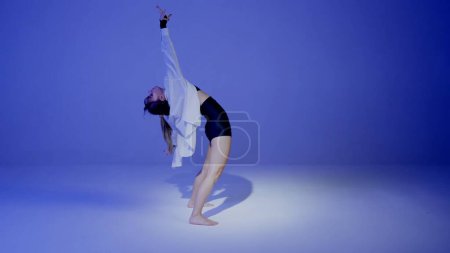 Photo for Young woman wearing a top, shorts and a shirt performing emotional contemporary dance under a spotlight in the studio. Neon blue color scheme, ombre, gradient background. Full length. - Royalty Free Image