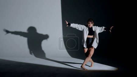 Photo for Young woman wearing a top, shorts and a shirt performing emotional contemporary dance under a spotlight in the studio. Contrasting black falling shadow on the background. Full length. - Royalty Free Image