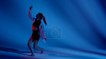 Photo for Young woman wearing a top, shorts and a shirt performing emotional contemporary dance in studio. Neon blue and red color scheme, shadowed background. Full length. Advertisement, creative content. - Royalty Free Image