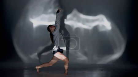 Photo for Young woman wearing a top, shorts and a shirt performing emotional contemporary dance under the glare of the light in the studio. Contrasting black falling shadow on the background. Full length. - Royalty Free Image