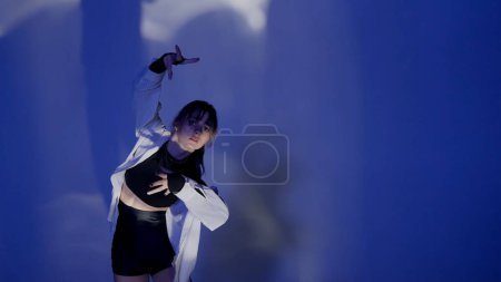 Foto de Mujer joven vestida con un top, pantalones cortos y una camisa realizando emotiva danza contemporánea bajo el resplandor de la luz en el estudio. Esquema de color azul neón, fondo sombreado. Medio lleno. - Imagen libre de derechos