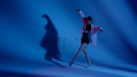 Foto de Mujer joven vistiendo un top, pantalones cortos y una camisa realizando danza contemporánea emocional en el estudio. Esquema de color azul y rojo neón, fondo sombreado. Longitud total. Publicidad, contenido creativo. - Imagen libre de derechos