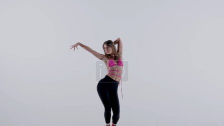 Foto de Mujer joven que lleva un sujetador de color rosa brillante, la parte superior, leggings y tacones altos realizando baile tacones enérgicos o posando en el estudio, fondo blanco aislado. Longitud total. Publicidad, contenido creativo. - Imagen libre de derechos