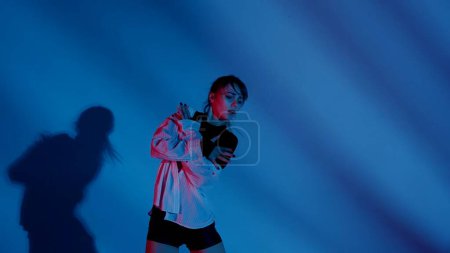 Photo for Young woman wearing a top, shorts and a shirt performing emotional contemporary dance in studio. Neon blue, pink and red color scheme, shadowed background. Medium sized. Advertisement, creative - Royalty Free Image
