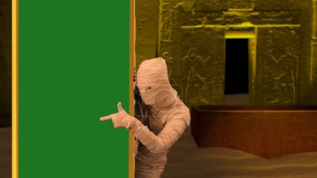 Photo for Egyptian pharaoh entombment. Scary a mummy peeking from behind a column with a green screen banner and pointing at it. Mock up, workspace for your promotion clip or advertisement. Halloween holidays. - Royalty Free Image