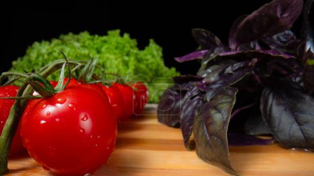 Photo for Macro shot of ripe juicy tomatoes, lettuce and basil sprigs in water drops. Fresh vegetables and herbs lie on a wooden table on a black background. Ingredients for a healthy diet salad - Royalty Free Image