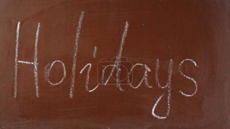 Photo for Textured brown chalkboard background. Holidays is written on the board with a piece of white chalk. Close up shot. Creative or educational content. - Royalty Free Image