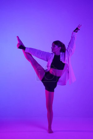 Foto de Mujer joven con un top, pantalones cortos y una camisa realizando danza contemporánea en el estudio. Esquema de color rosa neón y púrpura, ombre, fondo degradado. Longitud completa. - Imagen libre de derechos