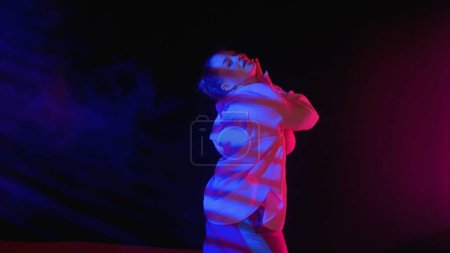 Téléchargez les photos : Belle femme dansant du jazz-funk dans un studio. Fond noir rempli de fumée, lumière rose et bleue et ombre tombante rayée. Chorégraphie moderne. Moyen plein. Clip promotionnel ou publicité. - en image libre de droit