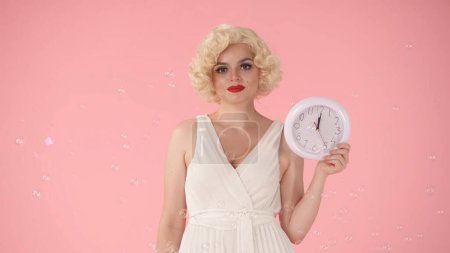 Photo for Woman holding a white round wall clock in her hand. Woman in image on Marilyn Monroe, in studio surrounded by soap bubbles on pink background. Concept of time - Royalty Free Image