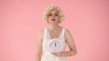 Foto de Mujer sosteniendo un reloj redondo blanco en la mano. Mujer en imagen sobre Marilyn Monroe, en estudio rodeada de pompas de jabón sobre fondo rosa. Concepto de tiempo - Imagen libre de derechos