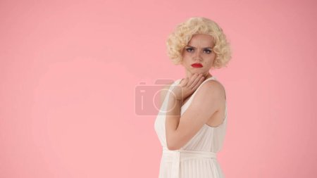 Foto de Vista lateral de una joven ofendida con labios pucheros. Retrato del perfil de la mujer que se parece a Marilyn Monroe, con vestido blanco y peluca blanca en el estudio sobre fondo rosa - Imagen libre de derechos