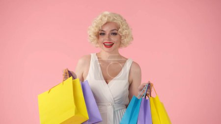 Téléchargez les photos : Jeune femme heureuse tenant des sacs à provisions colorés. Femme ressemblant à Marilyn Monroe en studio sur fond rose. Concept shopping, vente, Black Friday - en image libre de droit