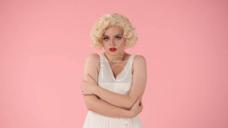 Foto de Una joven se siente ofendida. Retrato de una mujer en la imagen de Marilyn Monroe, vistiendo un vestido blanco y una peluca blanca en un estudio sobre un fondo rosa - Imagen libre de derechos