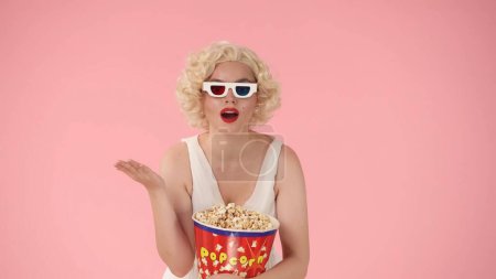 Foto de Mujer sorprendida, conmocionada con gafas 3d y sosteniendo un gran cubo de palomitas de maíz. Mujer en la mirada de Marilyn Monroe en el estudio sobre un fondo rosa - Imagen libre de derechos