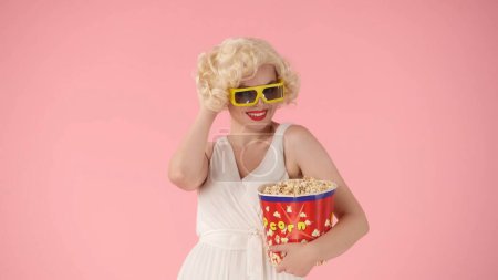 Foto de Mujer con gafas 3D y con un gran cubo de palomitas de maíz, disfrutando viendo una película. Mujer como Marilyn Monroe en estudio sobre fondo rosa - Imagen libre de derechos